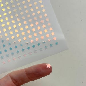 May include: Sheet of iridescent star stickers in various colors, including gold, pink, and blue. A single pink star sticker rests on a fingertip, showcasing the product's size and detail. The stickers are likely for crafts or decoration.