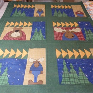 May include: Green fabric with a repeating pattern of moose and bears in a woodland setting. The fabric features a blue background with white stars and yellow triangles. The moose and bears are wearing plaid shirts and have brown fur.