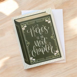 Op de afbeelding: Een wenskaart met een groen boekontwerp. De kaft heeft de tekst "Here's to the next chapter" in witte letters, met bloemen en bladeren. De kaart ligt op een gestreepte achtergrond.