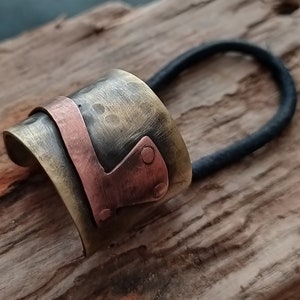 May include: A hair accessory featuring a wide, curved, hammered brass cuff with a polished copper accent piece attached by two small rivets. A black elastic hair tie is threaded through the metal cuff, creating a functional hair band design.