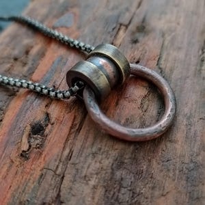 Op de afbeelding: Een handgemaakte ketting met een koperen ringhanger en een cilindrische bronzen kraal. De hanger hangt aan een donkere ketting. De ketting is geplaatst tegen een rustieke houten achtergrond, die de texturen en tinten van het metaal benadrukt.