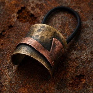 May include: A hair tie featuring a curved, cylindrical metal piece with a distressed, antiqued brass finish. A smooth, flat copper-colored metal band wraps around the center of the cylinder, secured by small rivets on one side of the hair accessory.
