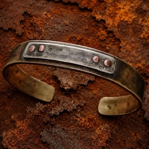 Op de afbeelding: Een brede, open armband van donker, verweerd metaal met een geborstelde afwerking. Het middenstuk bevat een rechthoekige plaat die is vastgezet met vier kleine, ronde koperkleurige klinknagels, met een kleine stempel aan de rechterkant.