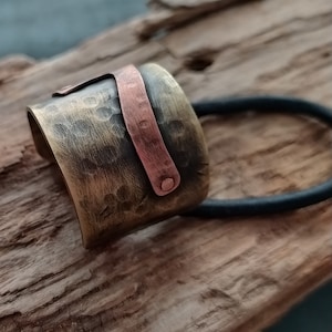 May include: A wide, cylindrical hair accessory featuring a hammered, antiqued brass finish. A smooth, contrasting copper-colored metal strip is riveted across the front surface. A black elastic hair tie is attached to the back of the metal piece.