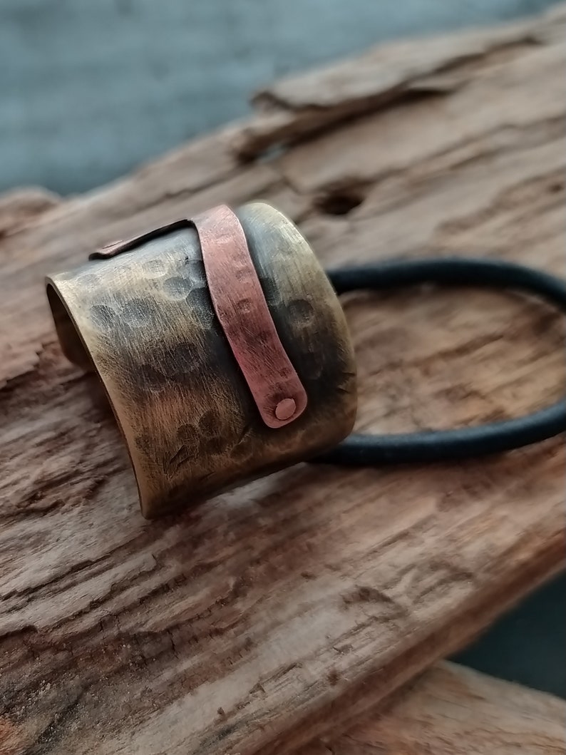 May include: A wide, cylindrical hair accessory featuring a hammered, antiqued brass finish. A smooth, contrasting copper-colored metal strip is riveted across the front surface. A black elastic hair tie is attached to the back of the metal piece.