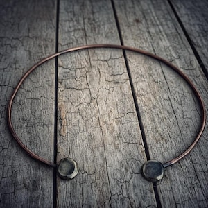 Op de afbeelding: Een koperkleurige choker-ketting met twee cirkelvormige, donkergrijze accenten. De ketting heeft een eenvoudig, minimalistisch ontwerp en ligt op een verweerd houten oppervlak. Het metaal heeft een licht verouderd uiterlijk.
