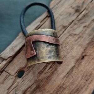 May include: A hair tie featuring a cylindrical metal cuff with a hammered brass finish and a contrasting horizontal copper band secured with small rivets. The cuff is attached to a thick, black elastic cord for securing hair in a ponytail.