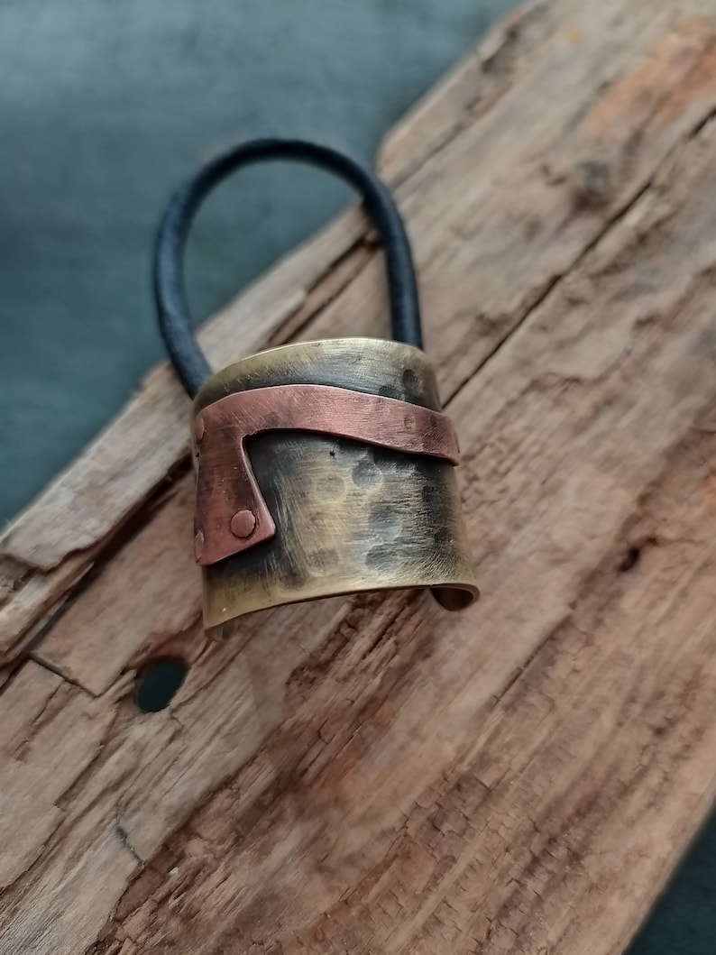 May include: A hair tie featuring a cylindrical metal cuff with a hammered brass finish and a contrasting horizontal copper band secured with small rivets. The cuff is attached to a thick, black elastic cord for securing hair in a ponytail.