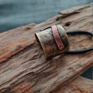 May include: A wide, hammered brass hair cuff featuring a contrasting vertical copper strip secured with a small rivet. The metallic surface has a dark, antiqued finish with visible indentations, and a black elastic hair tie is attached to the piece.