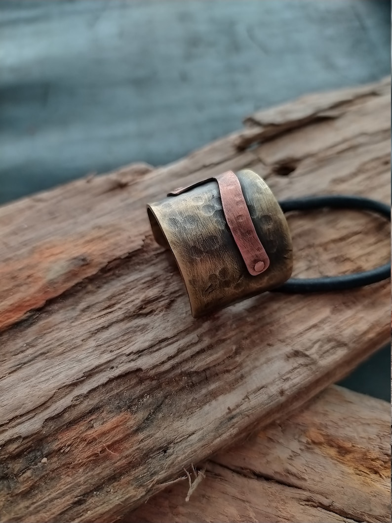 May include: A wide, hammered brass hair cuff featuring a contrasting vertical copper strip secured with a small rivet. The metallic surface has a dark, antiqued finish with visible indentations, and a black elastic hair tie is attached to the piece.