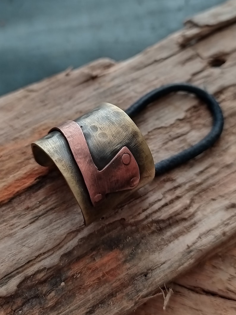 May include: A hair accessory featuring a wide, curved, hammered brass cuff with a polished copper accent piece attached by two small rivets. A black elastic hair tie is threaded through the metal cuff, creating a functional hair band design.