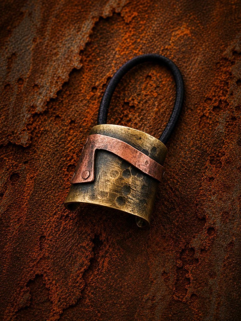 May include: A hair tie featuring a cylindrical brass cuff with a hammered, distressed finish. A curved copper strip is attached to the front of the brass cuff with a visible rivet. The elastic band is black and circular, attached to the metal component.