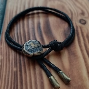 Puede incluir: Un brazalete de cordón negro con un dije de metal cuadrado y dos puntas de latón. El brazalete está atado con un nudo y tiene un extremo deshilachado.