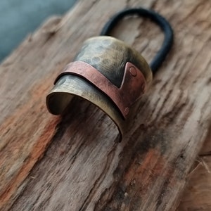 May include: A hair tie featuring a curved, hammered brass cuff with a dark, aged finish. A smooth, polished copper band wraps diagonally across the brass surface, secured by two small copper rivets on one side. The black elastic band is attached.