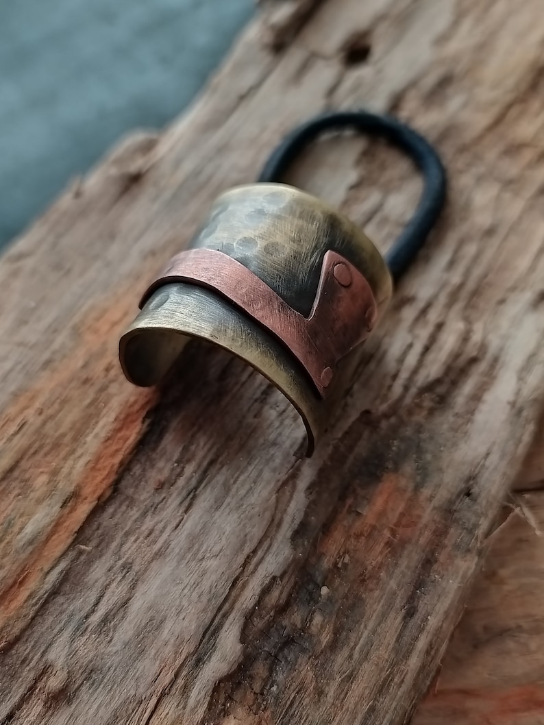 May include: A hair tie featuring a curved, hammered brass cuff with a dark, aged finish. A smooth, polished copper band wraps diagonally across the brass surface, secured by two small copper rivets on one side. The black elastic band is attached.