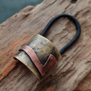 May include: A hair tie featuring a cylindrical metal cuff with a hammered, antiqued brass finish. A smooth, curved copper band wraps around the center of the cuff, secured by a small copper rivet on one side. The elastic band is black and circular.