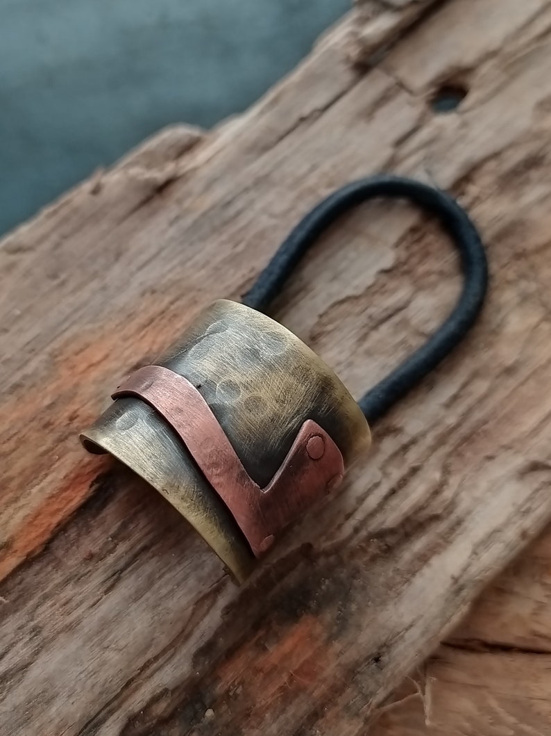 May include: A hair tie featuring a cylindrical metal cuff with a hammered, antiqued brass finish. A smooth, curved copper band wraps around the center of the cuff, secured by a small copper rivet on one side. The elastic band is black and circular.