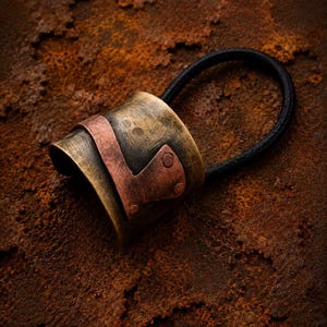May include: A hair tie featuring a cylindrical, hammered brass cuff with a weathered, antique finish. A contrasting, smooth copper-colored metal band is wrapped around the center of the brass cuff, secured by two small, visible rivets on one side.