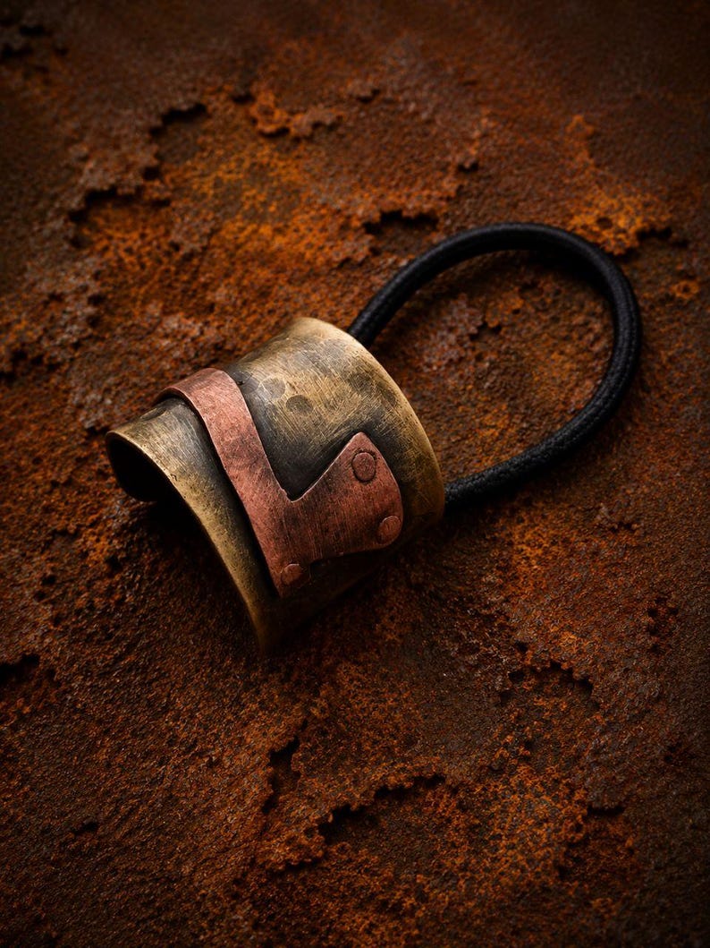 May include: A hair tie featuring a cylindrical, hammered brass cuff with a weathered, antique finish. A contrasting, smooth copper-colored metal band is wrapped around the center of the brass cuff, secured by two small, visible rivets on one side.