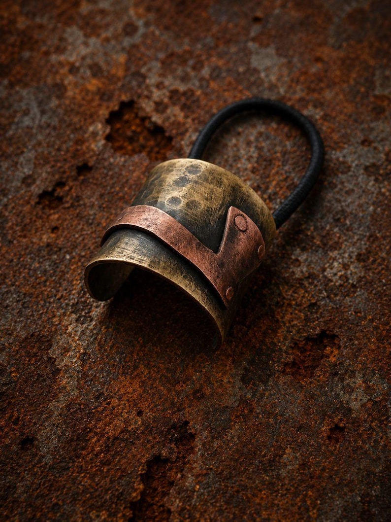 May include: A hair tie featuring a curved, cylindrical metal piece with a distressed, antiqued brass finish. A smooth, flat copper-colored metal band wraps around the center of the cylinder, secured by small rivets on one side of the hair accessory.