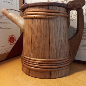 Könnte beinhalten: Ein Holzkrüg mit Henkel und Ausguss. Der Krug ist aus dunkelbraunem Holz mit hellbrauner Holzmaserung. Der Krug hat einen Band aus hellbraunem Holz um die Ober- und Unterseite. Der Krug steht auf einer hellbraunen Holzoberfläche. Der Krug trägt die Aufschrift "The Mary Rose Trust" auf der Seite.