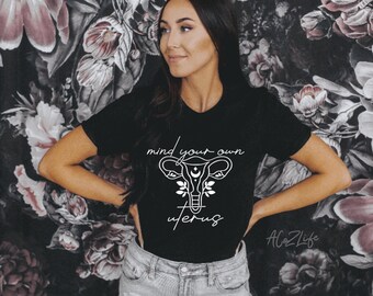 Peut inclure: T-shirt noir avec texte et graphiques blancs. Le texte dit "mind your own uterus" au-dessus d'un dessin stylisé d'utérus avec des éléments floraux et célestes. Le t-shirt est porté par une personne devant un fond floral.