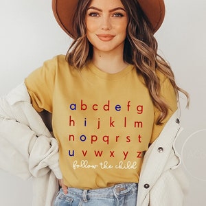 May include: A yellow t-shirt with the alphabet printed in red and blue. The letters are arranged in rows, with the phrase "follow the child" printed below the alphabet.