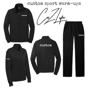 May include: Black custom sport warm-up set. The set includes a zip-up jacket and sweatpants. The jacket has a high collar and the word "custom" printed on the chest and sleeve. The sweatpants also have the word "custom" printed on the leg.