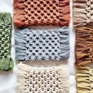 May include: A selection of handmade macrame coasters in shades of orange, green, grey, cream, and brown. Each coaster has a woven pattern with fringed edges. The coasters are displayed on a white surface.