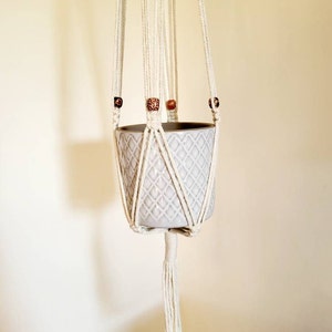 May include: A macramé plant hanger with a grey ceramic pot. The off-white cotton rope is knotted to hold the pot and features decorative wooden beads. The pot has a diamond pattern.