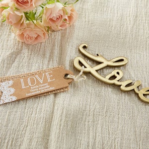 May include: A gold-colored bottle opener shaped like the word "LOVE" with a burlap and lace tag that reads "LOVE Bottle Opener, Ouvre-bouteille, Destapador de Botellas".