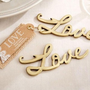 May include: Two gold-colored bottle openers shaped like the word "LOVE". The openers are on a white surface. One opener has a tag attached that says "LOVE Bottle Opener Ouvre-bouteille  Lechavador de Botellas".