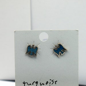 May include: A pair of silver-toned stud earrings with a turquoise inlay in a stylized bird design. The earrings are displayed on a white card with the words "turquoise inlay" printed on it. A ruler is at the bottom.