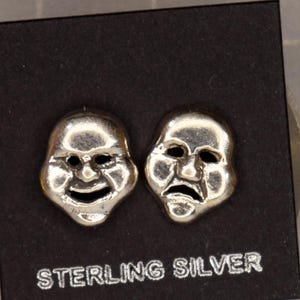 May include: A pair of silver-toned comedy and tragedy mask stud earrings mounted on a black display card with the text STERLING SILVER printed in white. A ruler on the right shows the earrings measure approximately 0.75 inches in width.