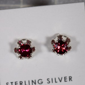May include: A pair of sterling silver stud earrings with a red gemstone in the center of each earring. The earrings are on a white card with the text "STERLING SILVER" printed on it.