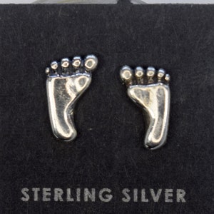 May include: Sterling silver stud earrings shaped like tiny footprints. The earrings are displayed on a black card with the words "STERLING SILVER" printed in white. The earrings are a bright silver color.