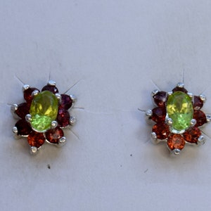 May include: A pair of stud earrings featuring a central oval-shaped, light green gemstone surrounded by a halo of smaller, deep red gemstones. The earrings have a silver-colored metal setting and a floral design.
