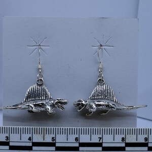 Könnte beinhalten: Ein Paar silberne Ohrringe mit einem detaillierten Dinosaurier-Design. Die Dinosaurier sind einander gegenübergestellt und etwa 3 cm lang.