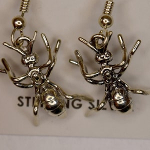 Könnte beinhalten: Ein Paar silberne Ameisenohrringe mit einem hängenden Design. Die Ohrringe befinden sich auf einem weißen Hintergrund mit einem Lineal im Hintergrund.