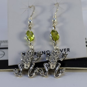 May include: Silver frog earrings with green gemstone accents. The earrings are on a white card with the text "Sterling Silver" printed on it.