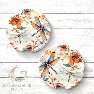 May include: Two round car coasters with a watercolor design featuring dragonflies and flowers in shades of orange, blue, and white. The coasters have a small indentation for easy removal from the car cup holder.