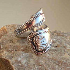 May include: A silver-toned wrap ring crafted from a repurposed spoon handle. The ring features a prominent embossed shell motif at the terminal end, with decorative scrollwork and linear detailing along the shank of the metal band.