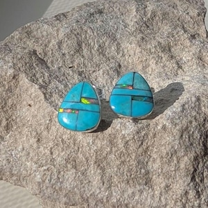 May include: A pair of teardrop-shaped stud earrings featuring inlaid turquoise segments separated by thin black lines, with small iridescent opal accents embedded within the stone patterns, set in polished silver metal frames.