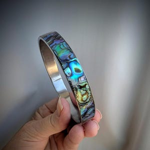 May include: A silver-toned bangle bracelet featuring an inlay of iridescent abalone shell. The shell displays a vibrant mosaic of blue, green, and purple hues with a polished finish. The metal band is smooth and solid, providing a clean contrast.