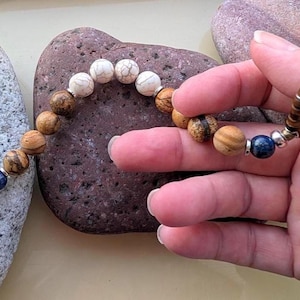 May include: A beaded bracelet featuring a mix of natural stone beads in shades of brown, white, and blue. The bracelet is held against a backdrop of textured stones, showcasing the earthy tones and unique patterns of the beads.