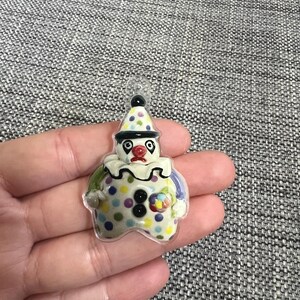 May include: A colorful clown-shaped pendant with a clear loop for hanging. The clown has a white face, a red nose, and a polka-dot hat and body. The pendant is held in a hand against a gray background.
