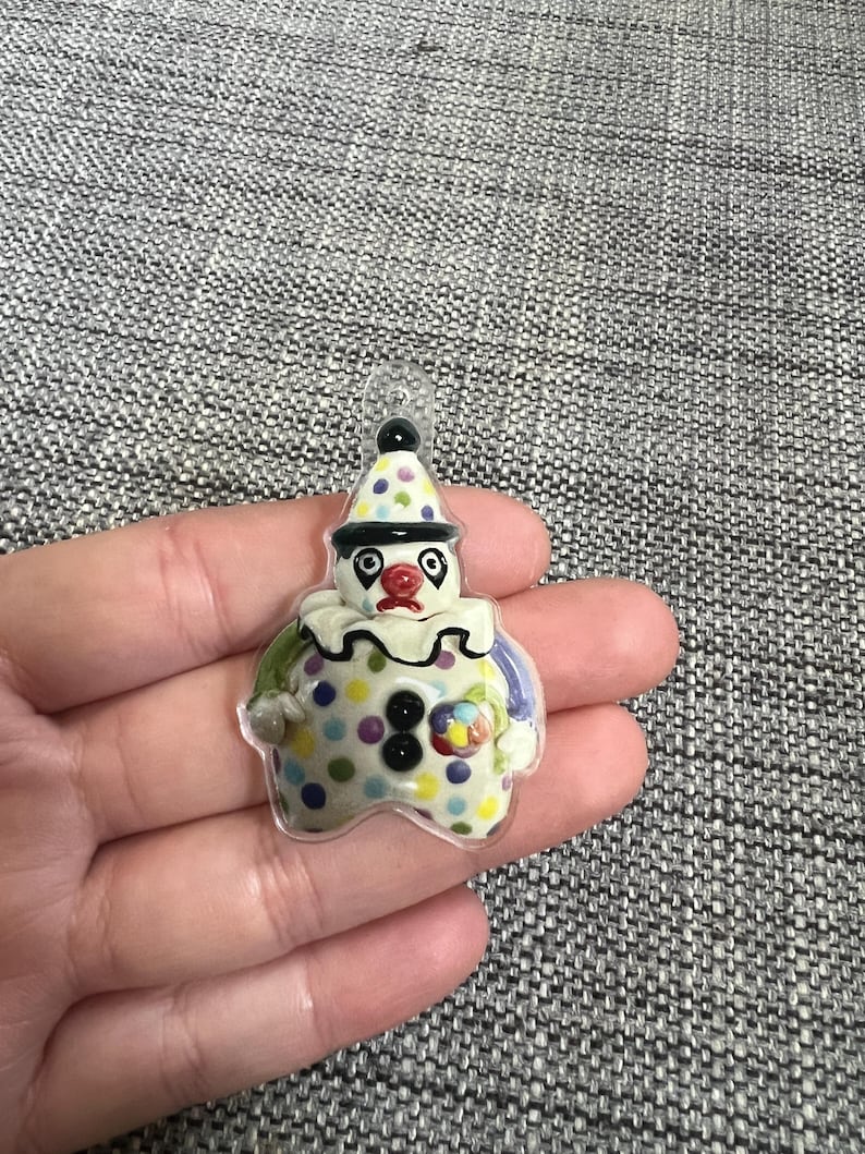 May include: A colorful clown-shaped pendant with a clear loop for hanging. The clown has a white face, a red nose, and a polka-dot hat and body. The pendant is held in a hand against a gray background.