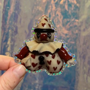 May include: A whimsical clown-shaped sticker with a holographic border. The clown has a cream-colored face, a red nose, and a black hat with red heart accents. The clown's body is cream-colored with red heart patterns.