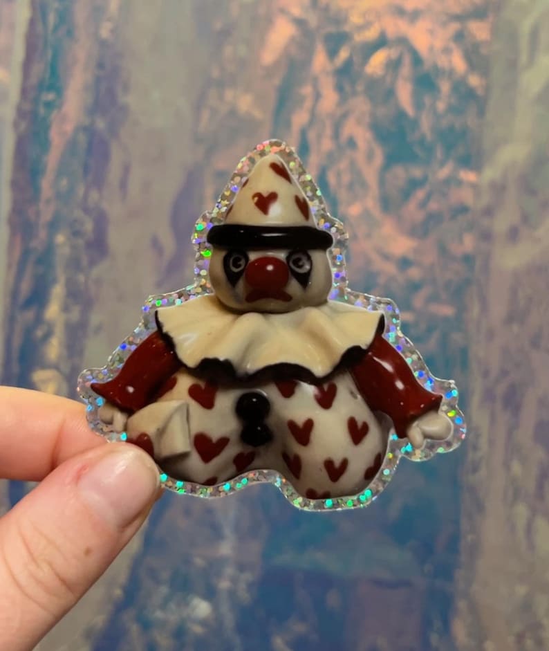 May include: A whimsical clown-shaped sticker with a holographic border. The clown has a cream-colored face, a red nose, and a black hat with red heart accents. The clown's body is cream-colored with red heart patterns.