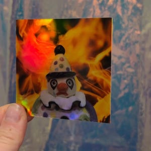 May include: A square sticker featuring a sad clown figurine with a polka-dot hat and ruffled collar. The background has a fiery, orange and yellow gradient. The sticker is held by a hand.